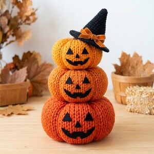 Könnte beinhalten: Ein Stapel aus drei orangefarbenen gestrickten Kürbissen mit schwarzen Jack-o'-Lantern-Gesichtern aus Filz. Der oberste Kürbis trägt einen schwarzen gehäkelten Hexenhut, der mit einer orangefarbenen Samtschleife verziert ist und eine Strickstruktur aufweist.
