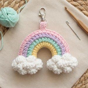 Könnte beinhalten: Ein gehäkelter Regenbogen-Schlüsselanhänger mit Bögen aus pastellrosa, lavendelfarbenem, mintgrünem und gelbem Garn. Der Regenbogen ist mit einem silbernen Karabinerhaken versehen und endet an beiden Seiten in flauschigen weißen Häkelwolken.