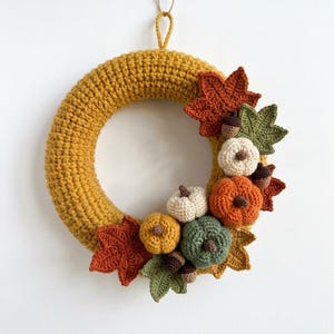 Può includere: Una ghirlanda circolare all'uncinetto color giallo senape, decorata con elementi autunnali lavorati all'uncinetto tra cui zucche arancioni, verdi e color crema, ghiande marroni e foglie d'acero nei toni ruggine, verde oliva e giallo oro.