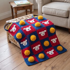 May include: A crochet blanket featuring a grid of alternating red and blue square motifs. Each square displays a white sheep face with black embroidered features. The blanket is accented with several bright yellow yarn pom-poms.