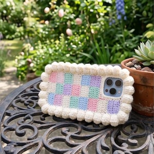 May include: A rectangular phone case featuring a crochet checkerboard pattern in pastel pink, mint green, light blue, and lavender, bordered by a thick, cream-colored yarn bobble trim. The case has a cutout for a triple-lens camera system.