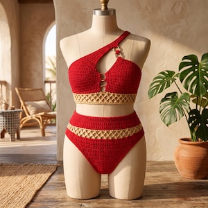 Puede incluir: Un conjunto de bikini de dos piezas tejido a ganchillo en rojo vibrante con ribete de patrón de celosía beige. La parte superior presenta un tirante asimétrico y detalles de anillos circulares dorados en el escote. La braga de talle alto hace juego.