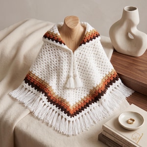 May include: A white crochet poncho featuring a hood and a V-shaped hem with long fringe. The garment is accented with horizontal stripes in shades of brown, tan, and orange near the collar and hem. Two white yarn tassels hang from the neckline.