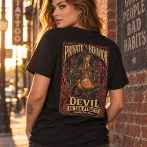Peut inclure: Un t-shirt noir avec une impression graphique au dos. Le motif représente un personnage central avec des cornes et une queue, entouré de roses rouges, de chaînes et de flammes. Le texte indique "PRIVATE BEHAVIOR", "DEVIL IN THE STREETS" et "Slightly Good News".