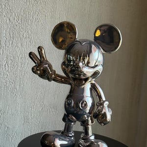 Puede incluir: Una figura de Mickey Mouse en tono plateado reflectante en pose de cuerpo completo. El personaje presenta grandes orejas redondas, una expresión de amplia sonrisa y una mano levantada en señal de paz. La superficie es pulida y metálica.