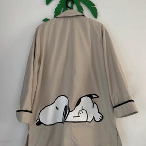 Könnte beinhalten: Ein beigefarbener Trenchcoat mit einem großen Grafikdruck der Zeichentrickfigur Snoopy, die auf der Rückseite liegt. Der Mantel hat lange Ärmel mit schwarzem Besatz an den Manschetten, einen klassischen Kragen und einen geraden Saum mit Seitenschlitzen.