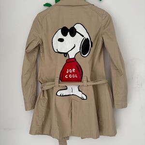 Può includere: Un trench beige con una grafica dipinta a mano sul retro che raffigura il personaggio dei cartoni animati Snoopy con occhiali da sole neri e un maglione rosso con la scritta JOE COOL in lettere bianche. Il cappotto è dotato di colletto e cintura.