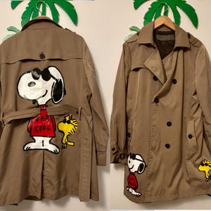 May include: A tan double-breasted trench coat featuring hand-painted illustrations of Snoopy and Woodstock wearing sunglasses. The back displays a large Snoopy in a red shirt with the text JOE COOL, while the front shows smaller versions.