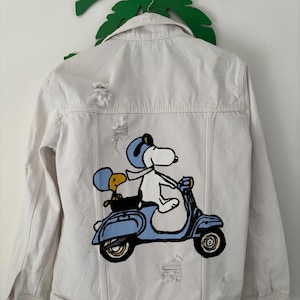May include: A white denim jacket featuring a distressed finish and a graphic print on the back. The illustration depicts a white dog wearing a blue helmet and goggles, riding a light blue scooter with a small yellow bird passenger sitting behind.