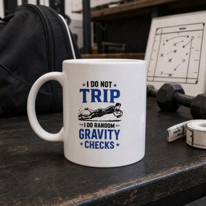 May include: A white ceramic mug featuring blue text that reads I DO NOT TRIP I DO RANDOM GRAVITY CHECKS. The design includes a black graphic of a person diving while holding a ball, positioned between the lines of text with small blue stars.