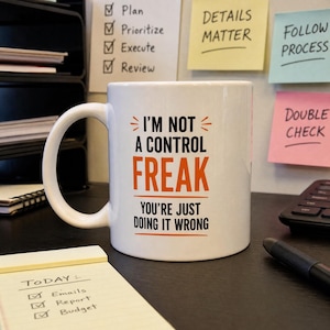 May include: A white ceramic mug featuring the black and orange text "I'M NOT A CONTROL FREAK YOU'RE JUST DOING IT WRONG" with orange horizontal lines above and below the word "FREAK" and small orange decorative marks on the sides.