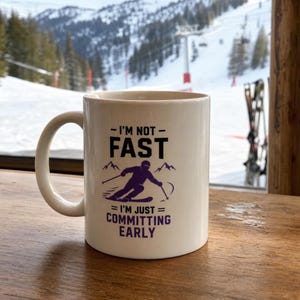 May include: A white ceramic mug featuring a purple graphic of a skier and the black and purple text: I'M NOT FAST I'M JUST COMMITTING EARLY. The mug has a curved handle and a smooth finish, displaying a clear design centred on the front surface.