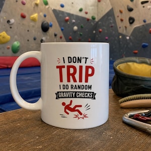 Può includere: Una tazza in ceramica bianca con il testo in nero e rosso I DON'T TRIP I DO RANDOM GRAVITY CHECKS e un grafico rosso di un omino stilizzato che cade. La tazza ha un manico curvo sul lato sinistro ed è posizionata in verticale.
