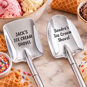 May include: Two polished stainless steel ice cream shovels with engraved text. The left shovel features the text JACK'S ICE CREAM SHOVEL, while the right shovel features the text Sandra's Ice Cream Shovel. Both have a spade-like head design.
