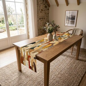 May include: A rectangular table runner featuring an abstract pattern of overlapping shapes in shades of brown, tan, cream, red, and black. The runner is laid flat across the centre of a wooden dining table, extending from one end to the other.