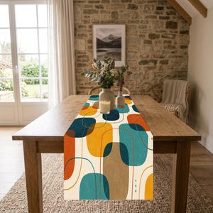May include: A rectangular table runner featuring an abstract pattern of rounded shapes in shades of orange, teal, dark blue, and beige on a cream background. The runner is centered on a wooden table with two vases and a small candle placed on top.