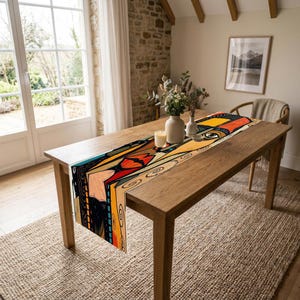 May include: A rectangular table runner featuring a vibrant, abstract graphic print with bold black, red, orange, and yellow geometric shapes and swirling line patterns. The design includes stylised facial features, such as an eye and lips, printed across the fabric, which is draped lengthwise over a wooden dining table.