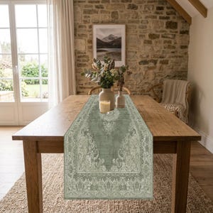 May include: A sage green table runner featuring an intricate, faded damask pattern with ornate floral and scrollwork motifs. The runner is centered on a rectangular wooden dining table, providing a decorative textile layer across the surface.
