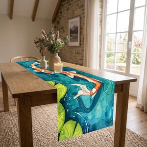 Può includere: Un runner da tavola rettangolare con un'illustrazione in stile acquerello di pesci koi arancioni e bianchi che nuotano in acqua blu profondo tra ninfee verde brillante. Il runner è posizionato su un tavolo in legno con un vaso e una candela.