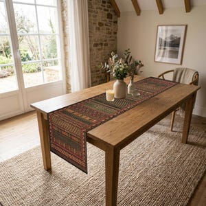 Puede incluir: Un camino de mesa rectangular con un patrón geométrico multicolor en tonos rojo, verde, naranja y negro. El camino está colocado sobre una mesa de comedor de madera de color marrón claro, centrado bajo un jarrón de cerámica blanca.