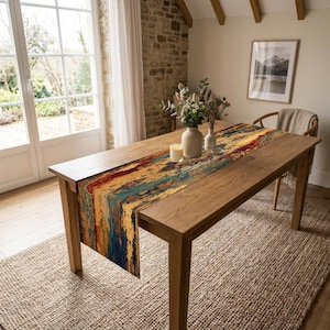 May include: A rectangular table runner featuring an abstract, multi-coloured pattern with deep blues, vibrant yellows, and rich reds. The runner is laid flat across a wooden dining table, accompanied by a white candle and a ceramic vase with greenery.