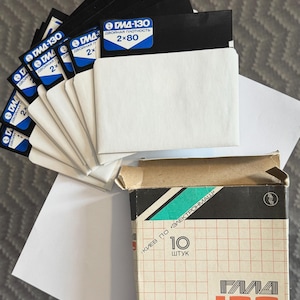 Könnte beinhalten: Ein Stapel schwarzer Disketten mit weißen Papierhüllen, jede mit einem blauen Etikett mit der Aufschrift GMD-130. Daneben steht ein offener Pappkarton mit Gittermuster, beschriftet mit dem Text 10 ШТУК und GMD-130 in Orange.