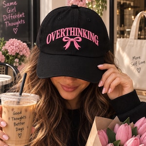 May include: A black baseball cap featuring the word OVERTHINKING embroidered in pink capital letters with a pink bow graphic centred below the text. The cap has a structured crown and a curved brim, designed with a classic six-panel construction.