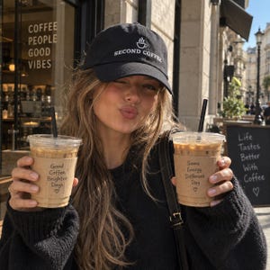 May include: A black baseball cap with the white embroidered text SECOND COFFEE and a small coffee cup icon. The cap features a curved brim and a structured crown. The text is centred on the front panel of the hat, which is made of a solid black fabric.