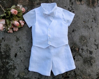 May include: White linen shorts set for a young boy, featuring a short-sleeved shirt, a vest with buttons, and a bow tie.