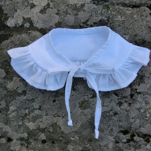 Op de afbeelding: Een witte kraag met ruches en een striksluiting. De kraag is gemaakt van een zachte stof en heeft een delicaat, vrouwelijk ontwerp. Een vintage-geïnspireerd accessoire.