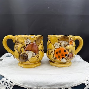 May include: Two yellow ceramic mugs featuring a raised, three-dimensional relief pattern of various mushrooms and small flowers. The mugs have a glossy finish, curved handles, and a scalloped rim, with one mug displaying a brown mushroom and the other an orange mushroom with black spots.