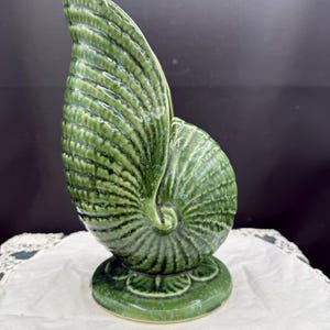 May include: A glossy, deep green ceramic vase shaped like a nautilus shell. The surface features intricate, raised radial ridges and a textured, spiraling pattern that culminates in a central point. It stands on a circular, pedestal-style base.