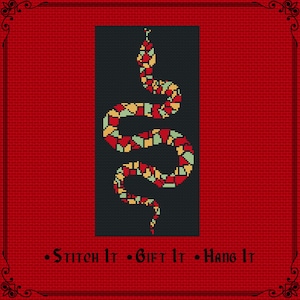 Può includere: Uno schema a punto croce raffigurante un serpente composto da blocchi geometrici rossi, gialli e verde chiaro su sfondo grigio scuro. Il testo "Stitch It • Gift It • Hang It" è stampato in nero in basso su un bordo rosso testurizzato.