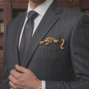 May include: A charcoal gray pinstripe blazer with a white collared shirt and a dark gray necktie. A gold-toned metal tiger brooch is attached to the breast pocket, featuring the tiger's head and front paws peeking out with its tail curled beside it.