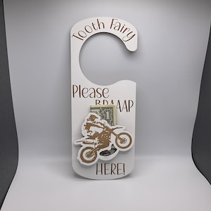 May include: White wooden door hanger with the text "Tooth Fairy Please Leave Here!" and a brown silhouette of a person riding a dirt bike. A dollar bill is tucked into the slot.