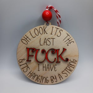 May include: A round wooden ornament with the text "Oh look it's the last f*** I have & it's hanging by a string" in black and red lettering. The ornament has a red and white string attached to it.