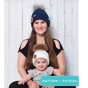 May include: A woman and a baby wearing matching knitted beanies with faux fur pom-poms. The woman's beanie is navy blue with wooden buttons, while the baby's is white. The text "PATTERN • PATRON" is visible.