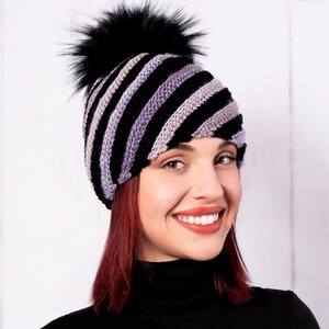 Puede incluir: Un gorro de punto de rayas negras y moradas con un pompón de piel sintética negra.