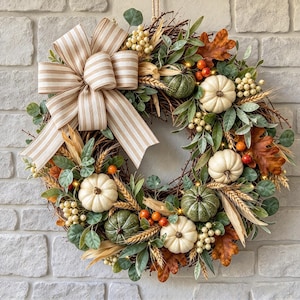 May include: A decorative wreath featuring a grapevine base adorned with white and green pumpkins, wheat stalks, brown oak leaves, and cream-colored berries. A large beige and white striped fabric bow is attached to the upper left side.