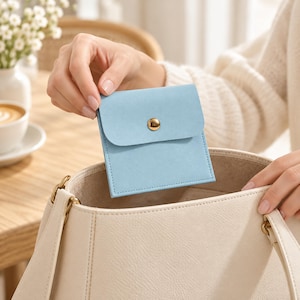 May include: A small, square, light blue pouch with a smooth texture and a single gold-toned metal snap button closure. The pouch is held above a large, off-white tote bag, featuring clean stitching along the edges and a flap top.