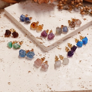 May include: A collection of raw gemstone stud earrings featuring various colors including blue, yellow, purple, green, and clear stones. Each earring is set in a gold-toned metal base, showcasing natural, irregular shapes and textures for each pair.