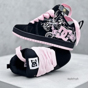 May include: A pair of black and pink DC sneakers featuring thick light pink laces, a black suede and patent leather upper with white swirling graphic patterns, a pink DC logo on the side, and a black sole with a pink trim. A cross charm is attached.