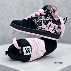 May include: A pair of black and light pink sneakers featuring a high-top silhouette, thick light pink laces, and a black side panel with a white floral-inspired graphic print. The DC logo is prominently displayed in light pink on the side.