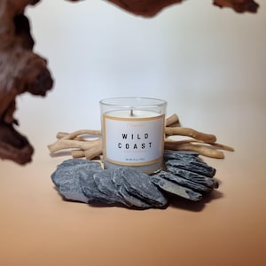 Peut inclure: Une bougie au soja dans un pot en verre transparent avec une étiquette blanche portant le texte Wild Coast et Net Wt. 6 oz / 170 g. La bougie est entourée de plusieurs pierres d'ardoise gris foncé plates et de brindilles en bois clair.