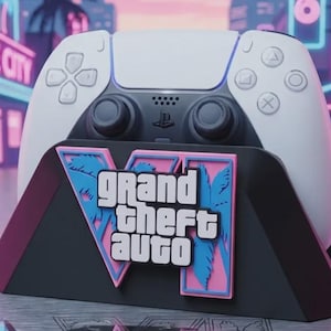 May include: A white and black video game controller resting in a black stand featuring a pink and blue Grand Theft Auto VI logo. The logo displays the text Grand Theft Auto with a large Roman numeral VI behind it, decorated with palm tree silhouettes.
