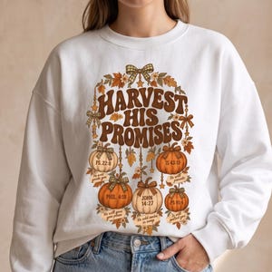 May include: A white crewneck sweatshirt featuring a graphic print with the text HARVEST HIS PROMISES. The design includes five pumpkins in shades of orange and cream, decorated with plaid bows, autumn leaves, and hanging stars.