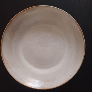 Może przedstawiać: Okrągła ceramiczna miska z matową, złamano-białą powierzchnią wewnętrzną i gładkim, lekko teksturowanym wykończeniem. Krawędź miski posiada cienki, ciepły brązowy brzeg, który kontrastuje z jaśniejszym środkiem, tworząc prosty, kolisty wzór.
