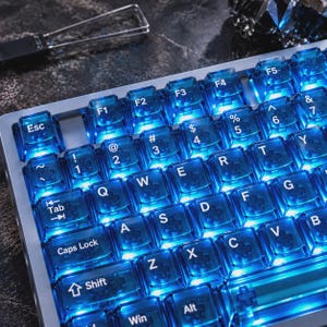 May include: A mechanical keyboard featuring translucent blue keycaps with white lettering and bright blue backlighting. The keys include standard QWERTY layout characters, function keys, and control keys like Esc, Tab, Caps Lock, Shift, Win, and Alt.