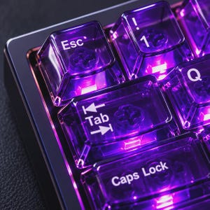 Puede incluir: Un teclado mecánico con teclas transparentes de color morado y letras blancas. Las teclas Esc, 1, Q, Tab y Caps Lock están iluminadas desde abajo por una luz morada brillante que resalta los mecanismos internos de los interruptores.