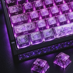 May include: A mechanical keyboard featuring translucent keycaps with embedded purple floral patterns and bright purple backlighting. The keyboard frame has a dark, glittery border, and two additional matching floral keycaps sit in the foreground.